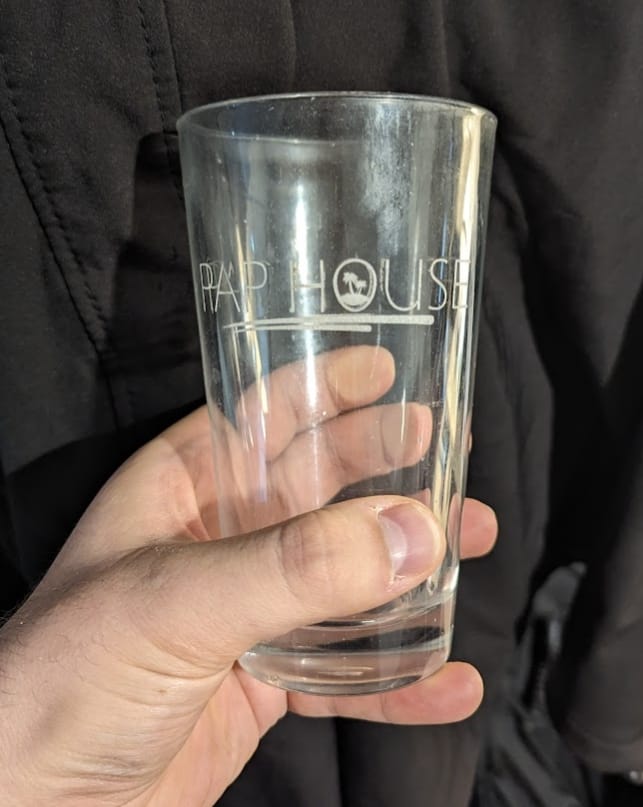 A hand holds a clear glass with "PAP HOUSE" etched on it, featuring a small palm tree logo.