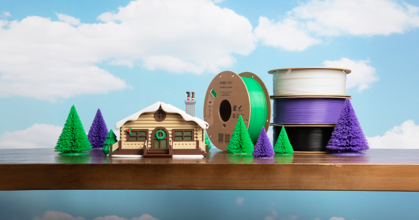 A detailed 3D printed winter cabin and colorful trees displayed on a wooden shelf next to spools of green, white, purple, and black filament.