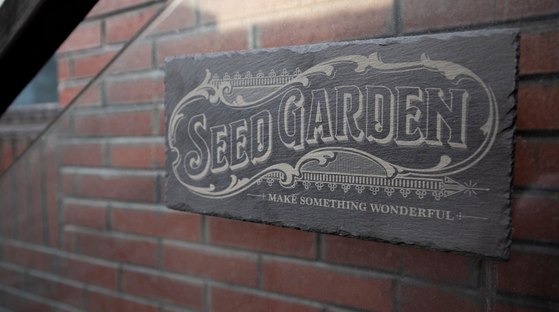 A finished laser-engraved slate sign with ornate "Seed Garden" lettering mounted on an exterior brick wall.