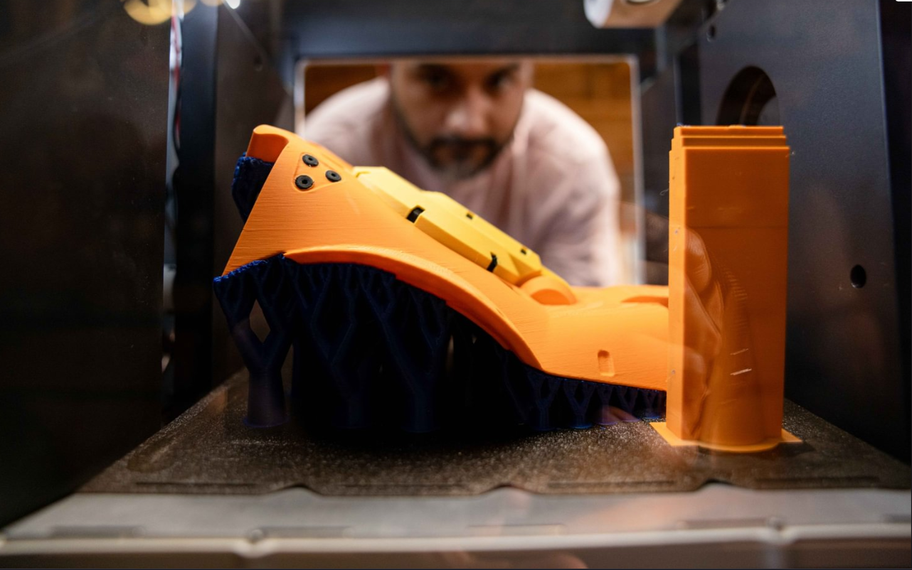 A view inside the printer enclosure showing an orange model on blue supports, featuring a rectangular purge tower on the right used to prime nozzles during material transitions.
