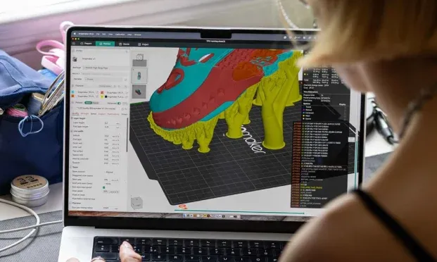 A designer using 3D slicer software on a laptop to generate yellow tree supports for a sneaker model before printing.