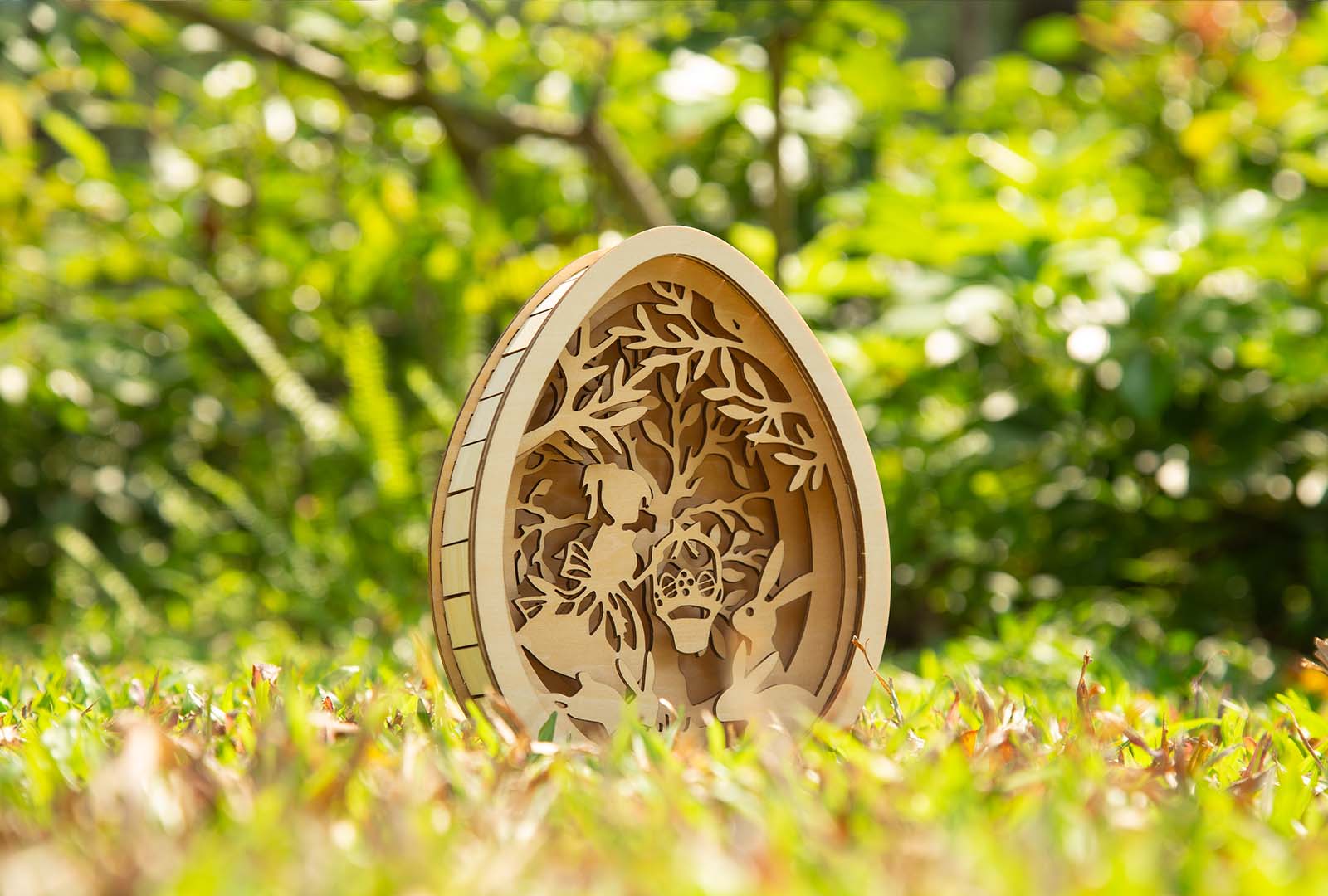 A multi-layered laser-cut wooden egg diorama depicting a detailed woodland scene with a rabbit and a figure, standing on sunlit grass.