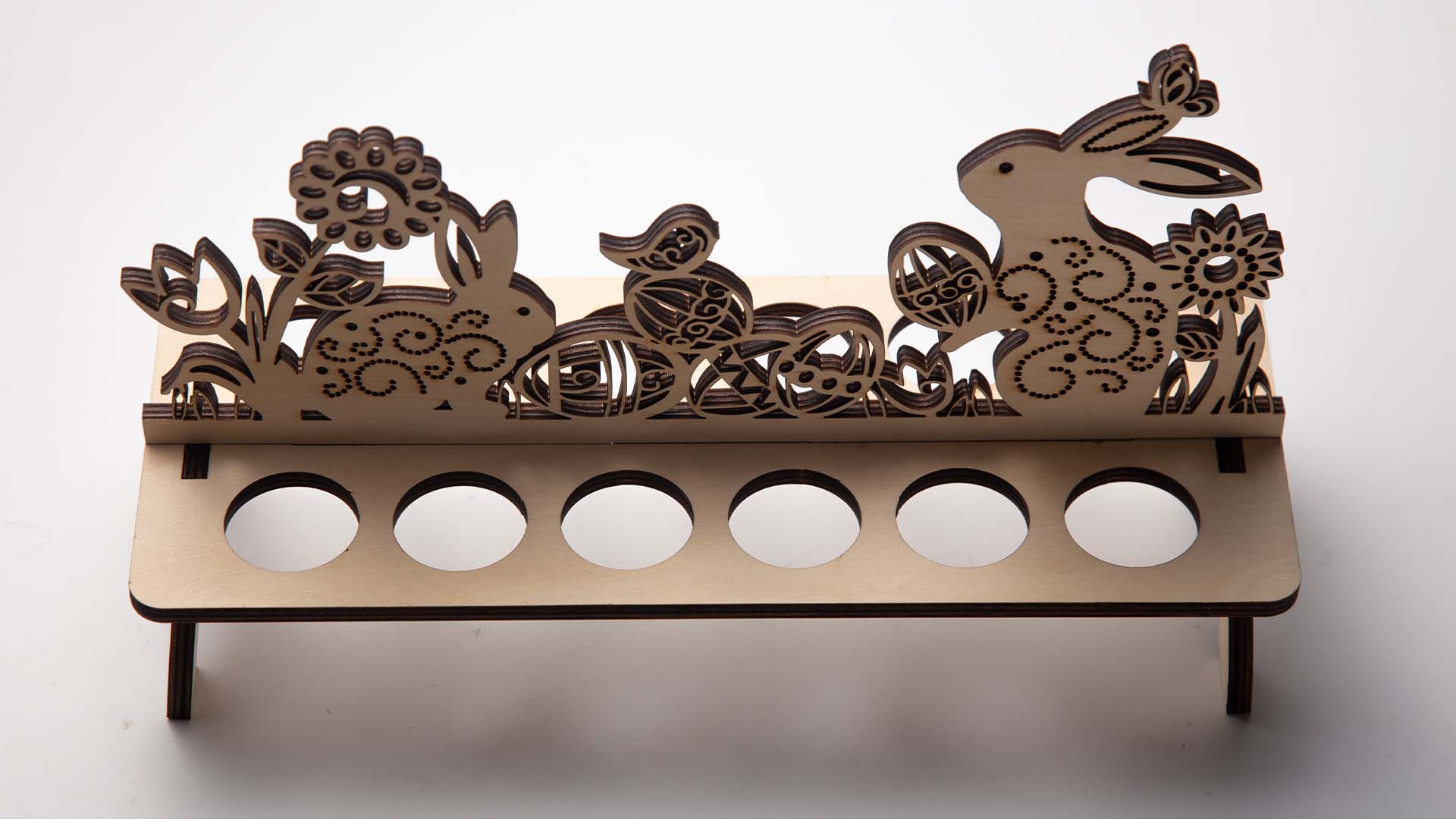 A detailed laser-cut wooden stand against a white background, featuring a scene with bunnies, plants, and eggs, and six circular holders designed to display decorated Easter eggs.