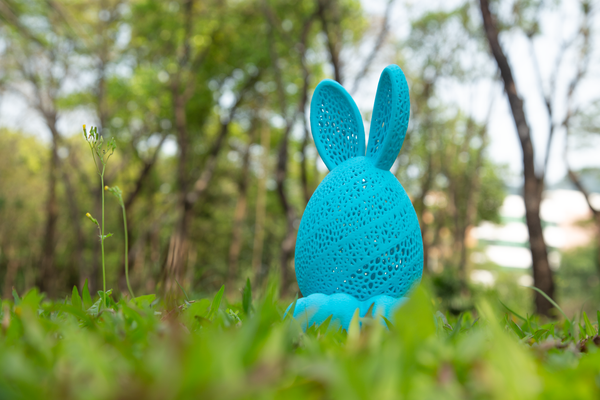 A hollow 3D printed blue bunny-shaped container with a porous Voronoi shell sitting in grass, illustrating how open-mesh designs eliminate the need for dense internal infill.