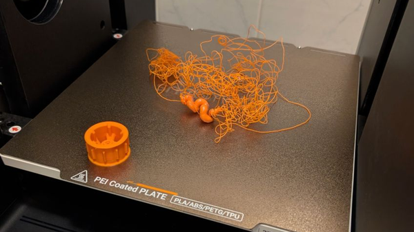 A failed 3D print on a PEI coated build plate showing a tangled "spaghetti" mess of orange filament, a common symptom of poor bed adhesion caused by incorrect nozzle height.